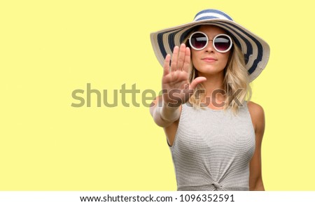 Young woman wearing sunglasses and summer hat annoyed with bad attitude making stop sign with hand, saying no, expressing security, defense or restriction, maybe pushing