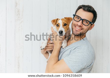 Horizontal portrait of handsome cheerful man, wears eyeglasses, holds jack russell terrirer, has glad expression, poses against white wooden wall with blank copy space. Animals and friendship Royalty-Free Stock Photo #1096358609
