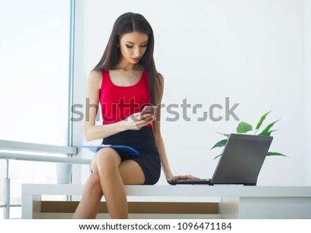 Business. Young Business Woman Sitting On The Table in the Big Light Office. Girl Dressed in Red T-Shirt and Black Skirt. Holding in Hand Folder. High Resolution