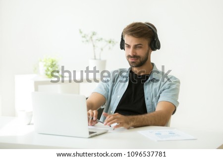 Concentrated Caucasian worker in headphones looking at laptop screen, watching webinar, listening to speaker, making notes at home or in office room. Male student taking online course, remote training