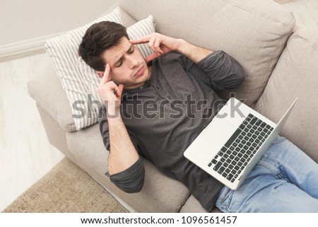 Unhappy tired man with laptop on couch at home office, suffering from headache, above view