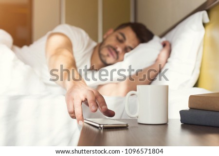 Sleepy guy waking up early after hearing alarm clock signal on smartphone on monday morning, reaching for ringing mobile phone with closed eyes, copy space Royalty-Free Stock Photo #1096571804