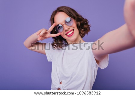 Pleasant girl with tattoo making selfie in studio and laughing. Good-looking young woman with brown wavy hair taking picture of herself on bright purple background.