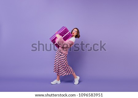 Lovable young woman in white sneakers posing with present box. Studio portrait of fascinating girl making kissing face expressing while holding big gift.