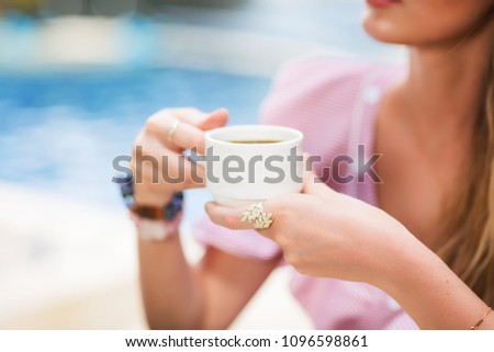 Outdoors lifestyle fashion image of young charming girl having breakfast. Holding in her hand cup of coffee. Drinking coffee, enjoying vacation. Wearing stylish silver rings, summer dress. Holiday 