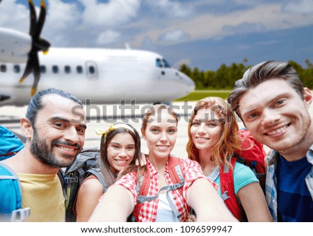 air travel, tourism and people concept - group of smiling friends with backpacks taking selfie over plane background