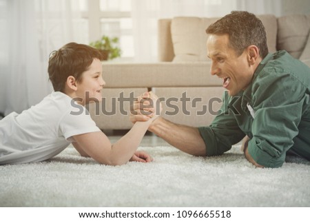 Lets see who is stronger. Cheerful father and son are doing arm-wrestling. They are lying on floor and laughing
