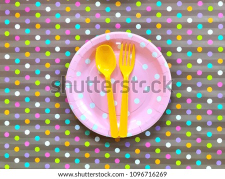 Closeup pale pink polka dot paper plate with yellow plastic spoon & fork on colorful polka dot background. Colorful disposable tableware for party,picnic.Top view with Copy space.Selective focus.