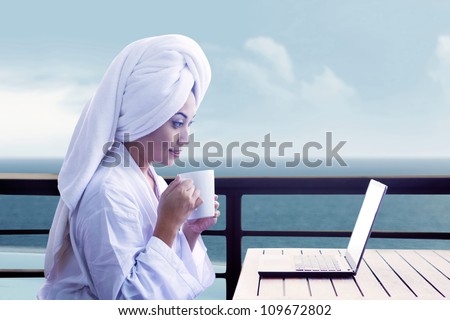 Asian woman in bathrobe holding cup of coffee and looking at laptop computer with ocean view