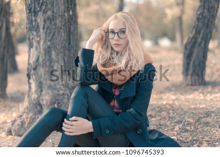 A girl in big black glasses sits park near a tree. Blonde in an orange woolen scarf and black coat look into the distance. On the ground lie yellow fallen leaves.