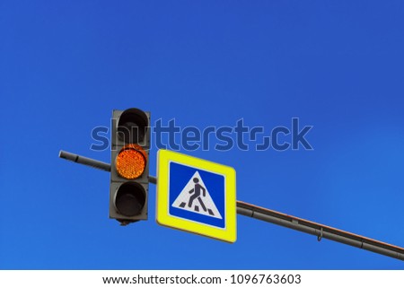 Yellow light traffic light signal and road sign of a pedestrian crossing on the console in the background of a bright sky with clouds. Copy space for text