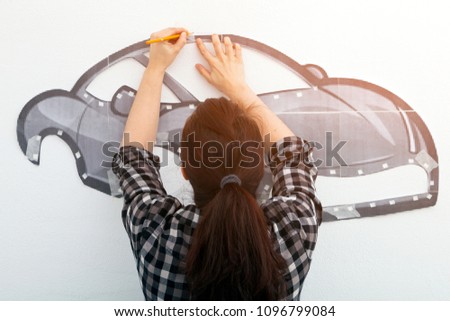 A young dark-haired woman in a plaid shirt draws a large red car in the boy's room on the wall. Mom paints his son car, he traces the contour of a law-book with a pencil