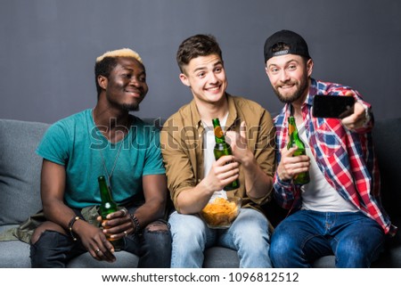 Portrait of stylish, best, attractive, modern positive guys with hairstyle, bristle sitting on couch, having mugs with lager in hands, shooting selfie on smart phone,