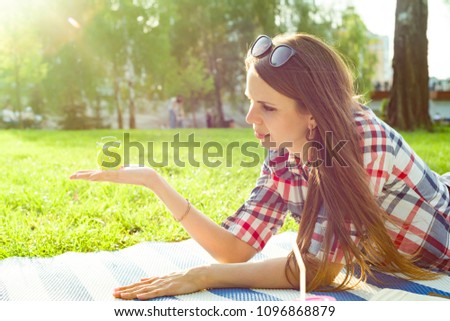 Young woman holding alarm clock in her hand, looking at the time. Background sunny city park, copy space