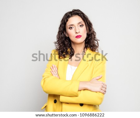 Studio portrait of confident young woman wearing yellow jacket, standing with arms crossed against grey background. 