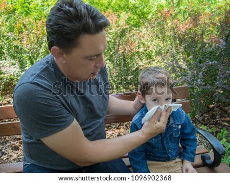 Father helps his son to blow his nose sitting on a bench in the park on a summer day with the green foliage of bushes around