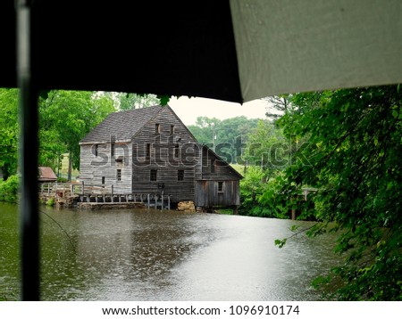 View of the old gristmill or watermill from across the pond on a rainy spring afternoon at Historic Yates Mill County Park in Raleigh North Carolina. Umbrella in frame.