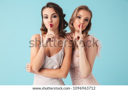 Shh! Image of two gorgeous women 20s wearing dresses and party makeup putting fingers on lips to keep secret isolated over blue background Royalty-Free Stock Photo #1096963712
