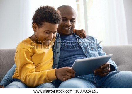 Interesting videos. Cheerful pre-teen boy hugging his father and watching videos together with him while relaxing on sofa