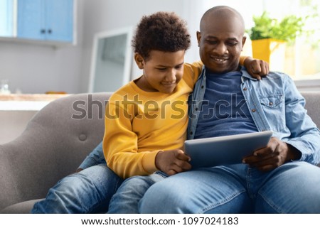 Funny videos. Pleasant young father and his pre-teen son sitting on the sofa and watching videos on tablet together while the boy hugging his father