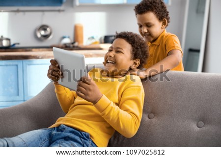 Brotherly care. Loving little boy giving his elder brother massage while the elder boy sitting on the sofa and playing a game on tablet