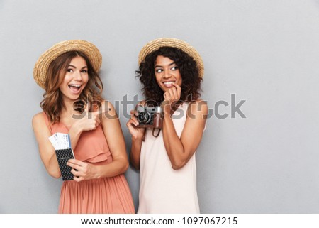 Portrait of two joyful young women dressed in summer clothes holding passport with tickets and photo camera over gray background