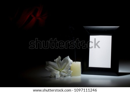 Blank thich black picture frame, with smoky candle and white lily flower on dark background with red decoration
