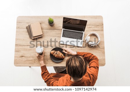 elevated view of man with coffee and croissant working at table with laptop, headphones, textbooks and potted plant