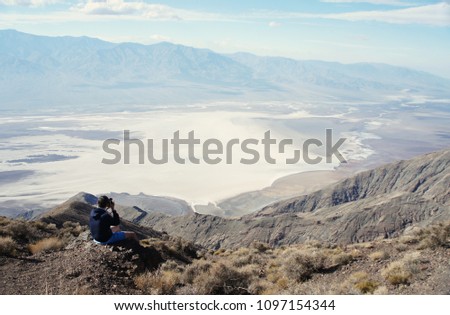 A photographer / videographer is sitting on the edge of the high rock overseeing and taking pictures of the fantastic landscape of Death Valley / USA. Copy space for the text. 