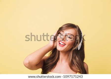 Photo of cute pleased woman 20s with long brown hair enjoying listen to music via headphones isolated over yellow background