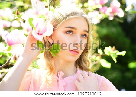 Pretty girl in pink blouse posing next to flowering tree. Blond young woman holding tender twig with gorgeous magnolia blossom. Relaxing walk on warm sunny day, spring time concept.