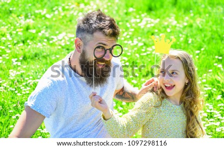 Dad and daughter sits on grass at grassplot, green background. Daddys princess concept. Family spend leisure outdoors. Child and father posing with crown and eyeglasses photo booth attributes