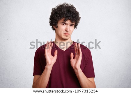Oh no, don`t bother me! Annoyed grumpy male with crisp hair, shows refusal gesture, poses against white background. Curly handsome teenager has dissatisfied expression, wears casual clothes. Royalty-Free Stock Photo #1097315234