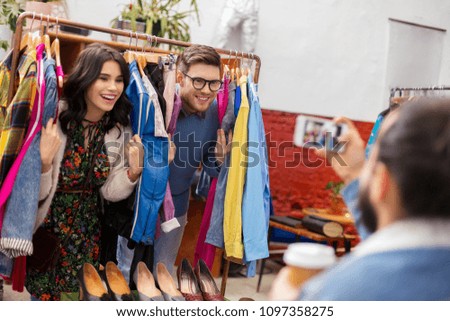 sale, shopping, fashion and people concept - friend photographing happy couple having fun by smartphone at vintage clothing store hanger