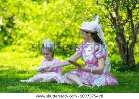 Family idyll, sitting on green grass in a sunny bright park in beautiful light vintage dresses in beads and hats, reading books and fooling around together. Mother and daughter in a summer park.