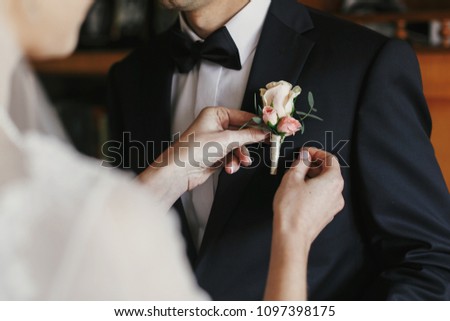beautiful bride putting on stylish simple boutonniere with roses on groom black suit. wedding morning preparations Royalty-Free Stock Photo #1097398175