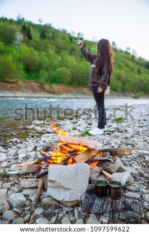 camping. young woman taking selfie while resting on riverside with camp fire