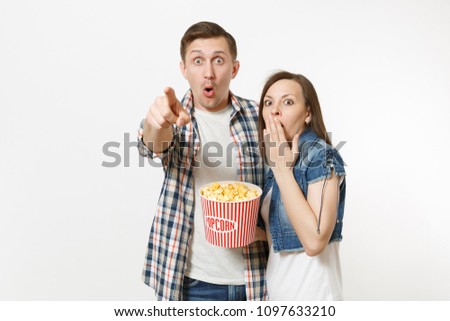 Young shocked couple, woman and man watching movie film on date, holding bucket of popcorn, pointing index finger on camera and covering mouth isolated on white background. Emotions in cinema concept