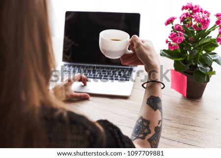 partial view of woman with tattooed hand holding cup of coffee and typing on laptop with blank screen at table