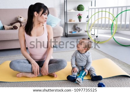 young beautiful mother and little child sitting on yoga mat at home and looking at each other