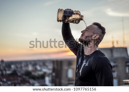 Young handsome tattooed man refreshing with water after training hard. Male sports outside on the top of the building. Outdoors recreation, stretching and training fit body. Royalty-Free Stock Photo #1097765156