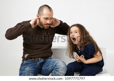 Angry little girl with long loose hair shouting, misbehaving because father didn't allow her watching cartoons. Frustrated young bearded man plugging ears, can't stand annoying screams by his daughter