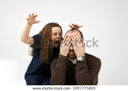 Fear me. Picture of funny European female kid dressed casually opening mouth widely, shouting, frightening her young stylish dad who is sitting and covering eyes, feeling scared and terrified