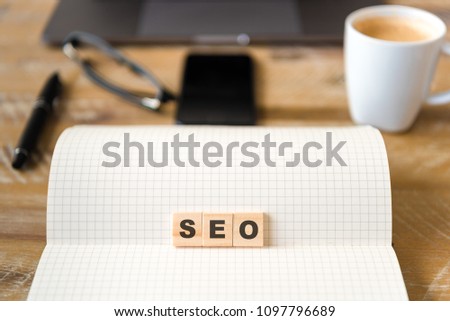 Closeup on notebook over wood table background, focus on wooden blocks with letters making SEO word. Business concept image. Laptop, glasses, pen and mobile phone in defocused background