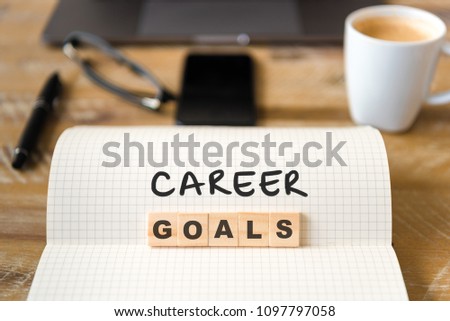 Closeup on notebook over wood table background, focus on wooden blocks with letters making Career Goals text. Concept image with laptop, glasses, pen and mobile phone in defocused background