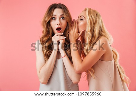 Pretty blonde woman in pajamas talking something to ear of shocked brunette woman over pink background Royalty-Free Stock Photo #1097855960