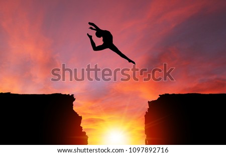 Silhouette of girl dancer in a split leap over dangerous cliffs with sunset or sunrise background and copy space. Concept of faith, conquering adversity, taking risk; challenge, courage, determination Royalty-Free Stock Photo #1097892716