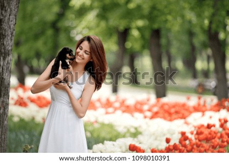 European GIRL IN A WHITE DRESS WITH DARK HAIR In the park with tulips hugs a small puppy