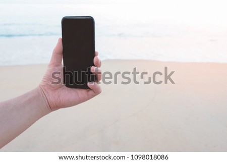 Close up man hand holding black smartphone on beautiful calm blue sea and sandy beach background. Male hand using mobile phone in outdoor summer beach in holiday vacation to taking picture.