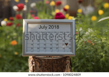 Calendar of the month July 2018 with lunar days & holidays is in a summer garden among blossom flowers. Parents' day is marked with a red heart.